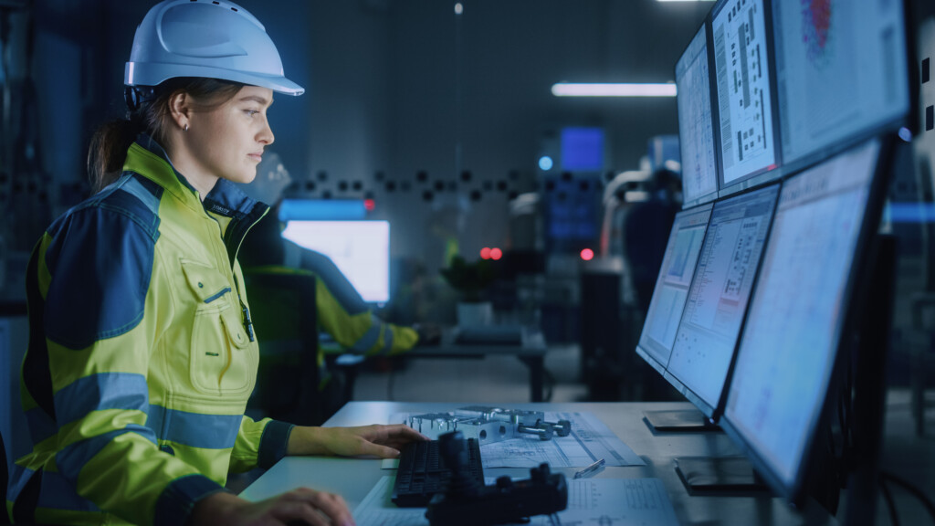 Industry 4.0 Modern Factory: Female Facility Operator Controls Workshop Production Line, Uses Computer with Screens Showing Complex UI of Machine Operation Processes, Controllers, Machinery Blueprints, Image by Adobe Stock